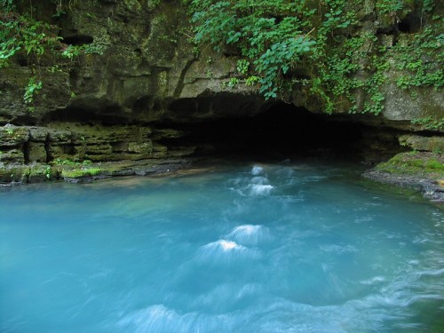 A serene blue river flows into a dark cave, surrounded by lush green foliage and rocky cliffs.