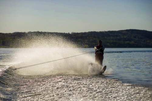 A person water skiing on a lake, creating a spray of water against a scenic backdrop of hills.