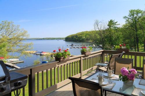 Scenic view of a lake from a deck with flowers, a grill, and seating, surrounded by lush greenery and clear blue skies.