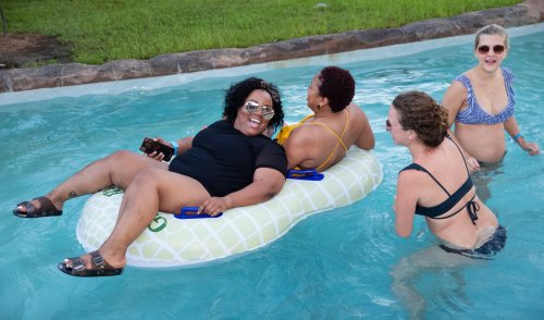 Four women enjoy a pool day, two on a float and two in the water, laughing and relaxing under the sun.
