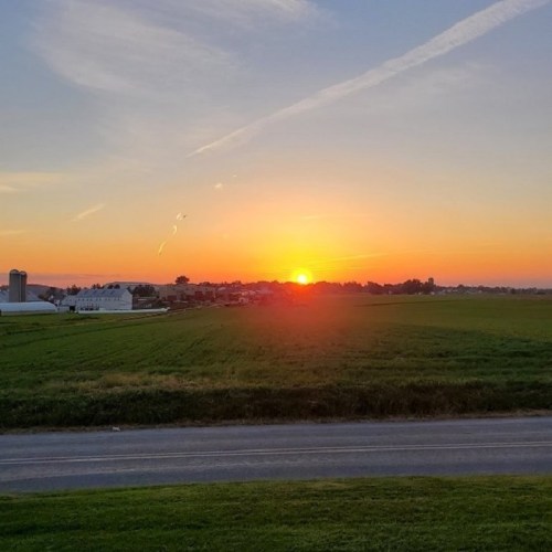 A vibrant sunset over a green field, with a distant farm and a clear sky streaked with clouds.