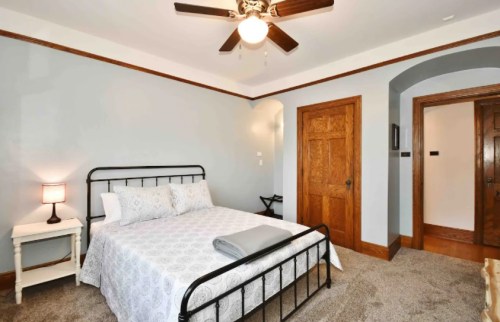 Cozy bedroom with a metal bed frame, gray walls, wooden accents, and a ceiling fan. Soft lighting from a bedside lamp.