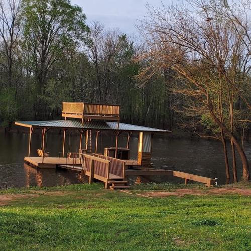 A wooden dock with a roof sits on a calm river, surrounded by trees and greenery.