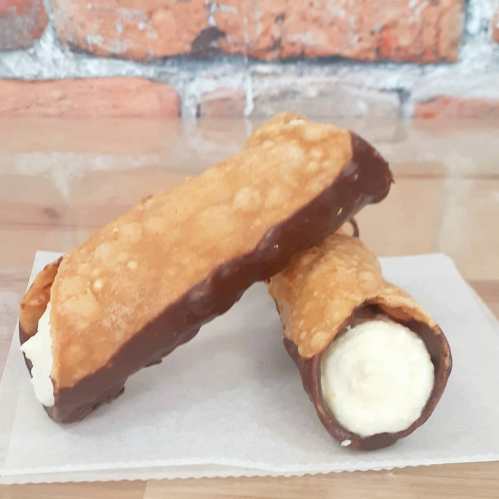 Two chocolate-dipped cannoli filled with creamy filling, resting on a piece of parchment paper against a brick background.