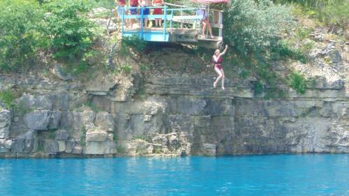 A person in a red swimsuit jumps off a cliff into a blue lake, with onlookers on a platform above.