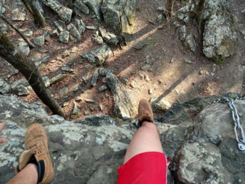 A person's legs in shorts and boots dangling over a rocky cliff, with trees and shadows in the background.