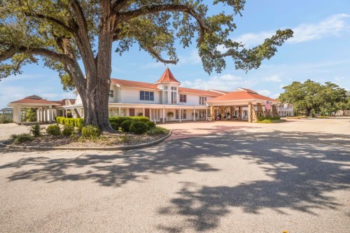 A large building with a red-tiled roof surrounded by trees and a paved driveway on a sunny day.
