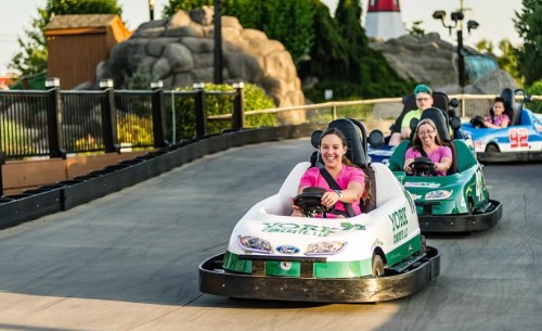 Two women in pink shirts drive go-karts on a racetrack, with a scenic background of trees and a lighthouse.
