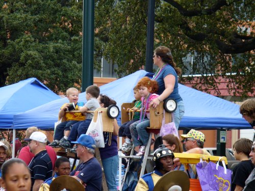 Children sitting on wooden platforms at a festive outdoor event, wearing beads and enjoying the lively atmosphere.