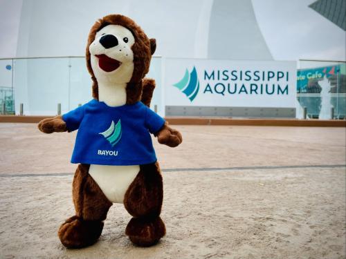 A plush otter wearing a blue "Bayou" shirt stands in front of the Mississippi Aquarium sign.