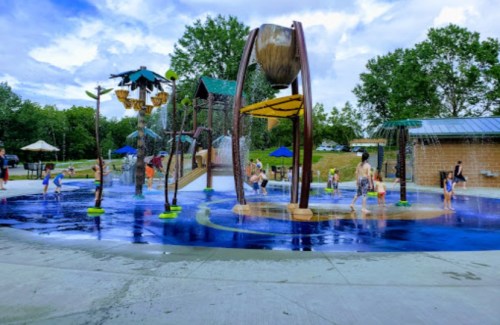 Children play in a colorful splash pad with water features, surrounded by trees and a cloudy sky.