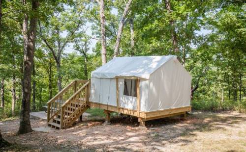 A white canvas tent on a wooden platform surrounded by trees, with a staircase leading up to the entrance.