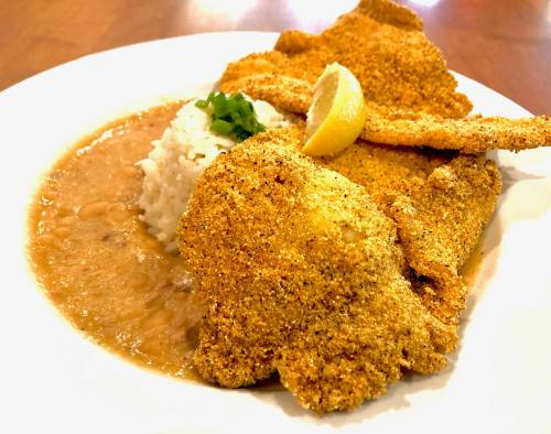 A plate of golden-fried fish fillets served with rice, beans, and a lemon wedge, garnished with cilantro.