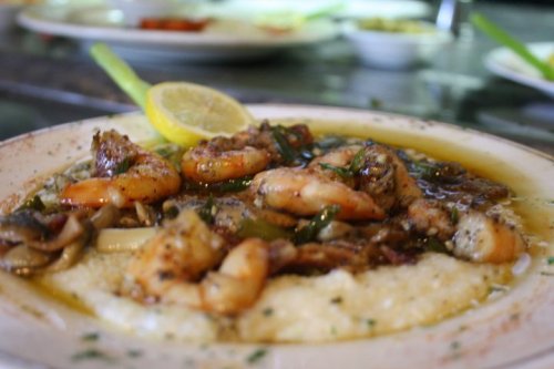 A plate of shrimp and grits topped with herbs, lemon, and a savory sauce, garnished with a celery stalk.