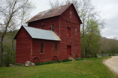 A red wooden building with a metal roof, surrounded by greenery and near a river, on a gravel path.