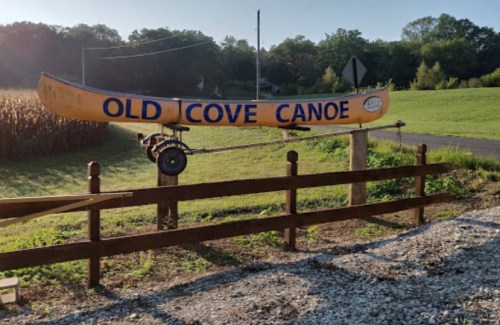 A large yellow canoe sign reading "Old Cove Canoe" mounted on a trailer near a grassy area and cornfield.