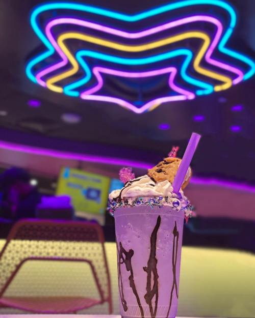A colorful milkshake topped with whipped cream and a cookie, set against vibrant neon lights in a modern café.
