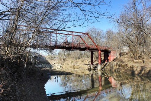 A red metal bridge spans a calm river, surrounded by bare trees and a clear blue sky. Reflections shimmer on the water.