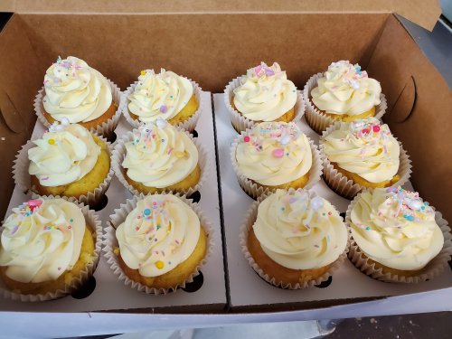 A box of twelve cupcakes with yellow frosting and colorful sprinkles, arranged in two rows.
