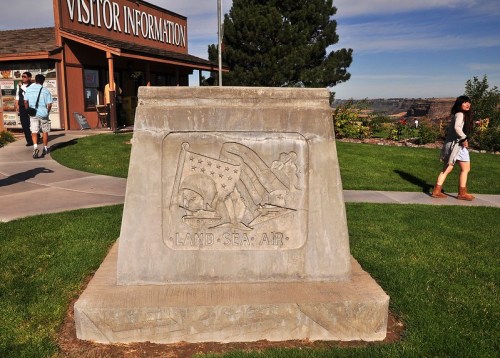 A stone monument with a relief carving, set in a grassy area near a visitor information center.