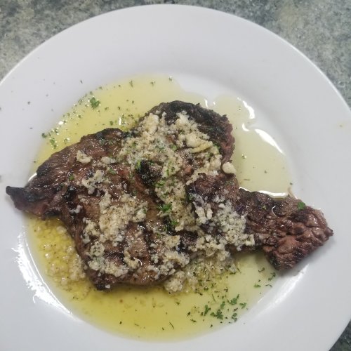 A grilled steak topped with crumbled cheese and herbs, served on a white plate with a drizzle of sauce.