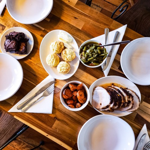 A wooden table set with plates of sliced turkey, green beans, carrots, cornbread, and brownies, with utensils nearby.