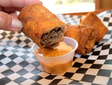 A hand holds a crispy egg roll, dipping it into a small cup of sauce, with more egg rolls on a checkered paper.