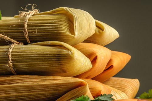 A close-up of stacked tamales wrapped in corn husks, featuring green and orange varieties, tied with twine.
