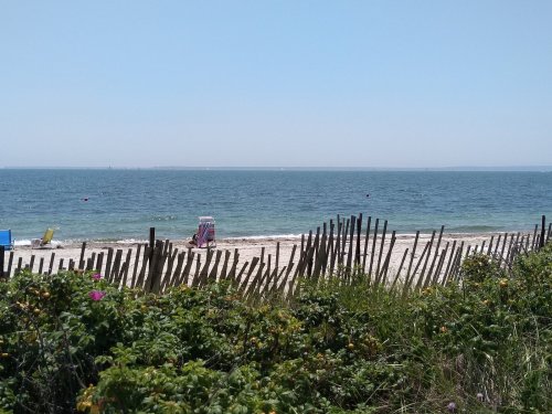 A serene beach scene with a sandy shore, gentle waves, and colorful beach chairs behind a wooden fence and greenery.