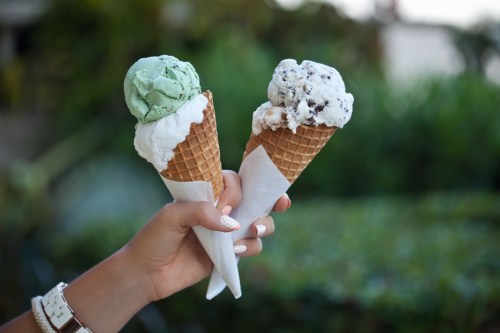 A hand holds two ice cream cones: one with mint green and the other with cookies and cream, against a blurred background.