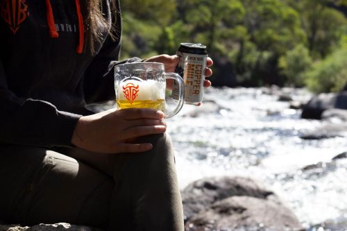 A person holds a glass of beer and a can by a river, enjoying a sunny day outdoors.