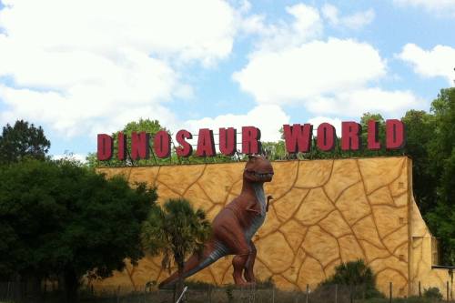 Sign for "Dinosaur World" with a large dinosaur statue in front, set against a blue sky and green trees.