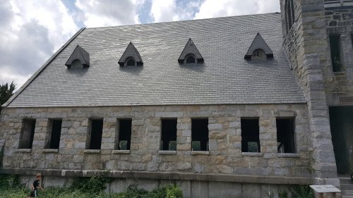 Stone building with a slate roof and arched windows, surrounded by greenery under a partly cloudy sky.