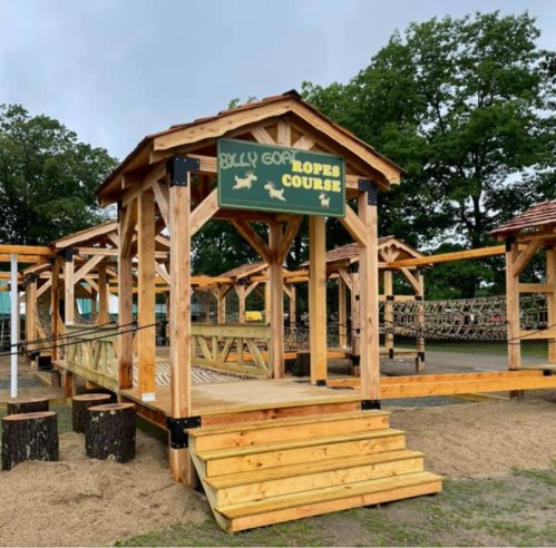 A wooden ropes course structure with a sign reading "Billy Goat Ropes Course" surrounded by trees and gravel.