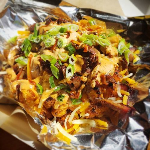 A foil tray filled with loaded nachos topped with grilled meat, cheese, green onions, and a creamy sauce.