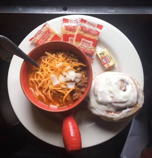 A bowl of chili topped with cheese and onions, served with crackers, butter, and a cinnamon roll on a plate.
