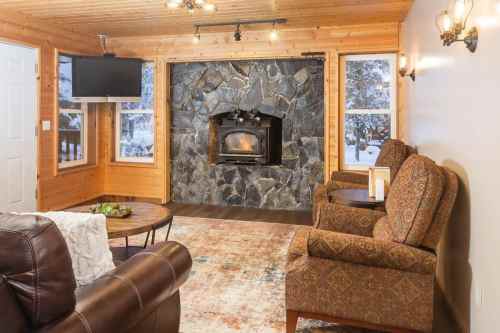 Cozy cabin interior with a stone fireplace, two armchairs, a coffee table, and a TV, surrounded by snowy views.