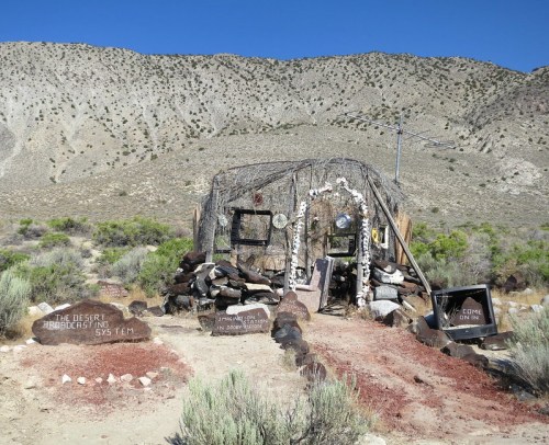 A rustic desert structure made of natural materials, surrounded by rocks and vegetation, with signs inviting visitors.
