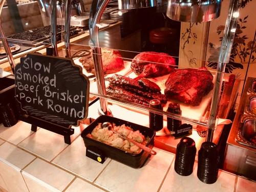 A display of slow smoked beef brisket, pork round, and sides at a barbecue restaurant, with a chalkboard sign.