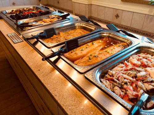 A buffet display featuring various seafood dishes in metal trays, including fish and crab, on a countertop.