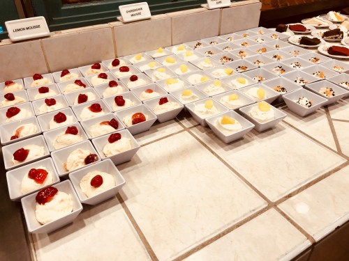 A variety of desserts in small bowls, including lemon mousse and cheesecake, arranged neatly on a tiled surface.