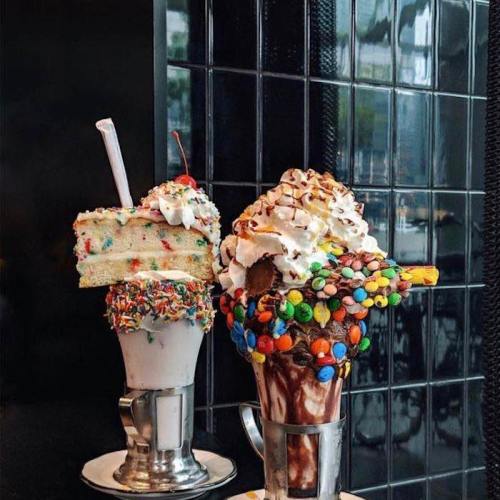 Two extravagant milkshakes topped with colorful sprinkles, whipped cream, and cake, served in metal cups.