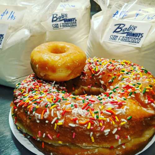 A large, colorful donut with sprinkles sits next to a smaller plain donut, with bags of flour in the background.