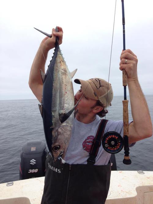 A fisherman on a boat holds a large fish close to his face, with a fishing rod in the other hand and water in the background.