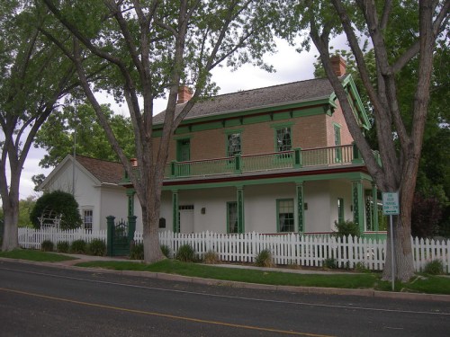 A two-story house with green trim, surrounded by trees and a white picket fence, located on a quiet street.