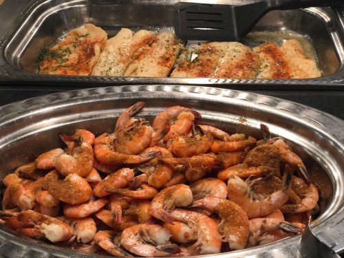 A silver serving tray with seasoned shrimp and a second tray with baked fish fillets, garnished with herbs.