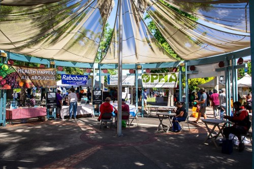 A vibrant market scene with banners promoting health, local vendors, and people enjoying the outdoor space under a canopy.