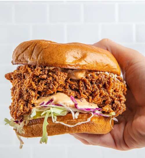 A hand holding a crispy fried chicken sandwich with lettuce and sauce on a soft bun.