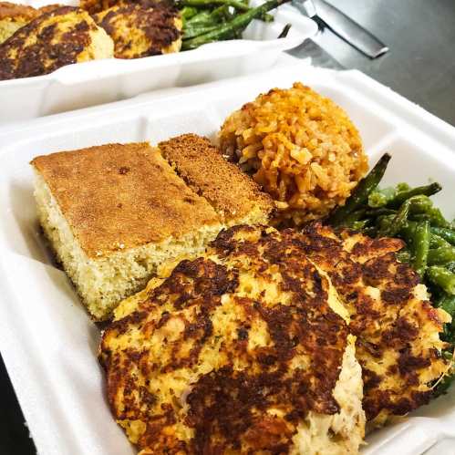 A takeout container with two crab cakes, green beans, cornbread, and a serving of rice.