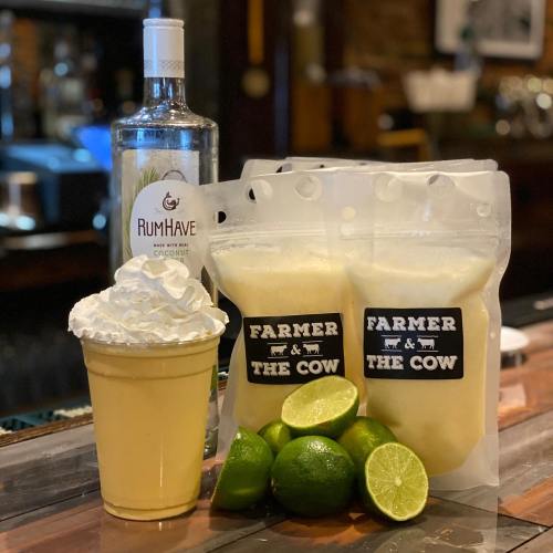 A creamy drink topped with whipped cream, surrounded by limes and pouches labeled "FARMER & THE COW," with a bottle of rum.